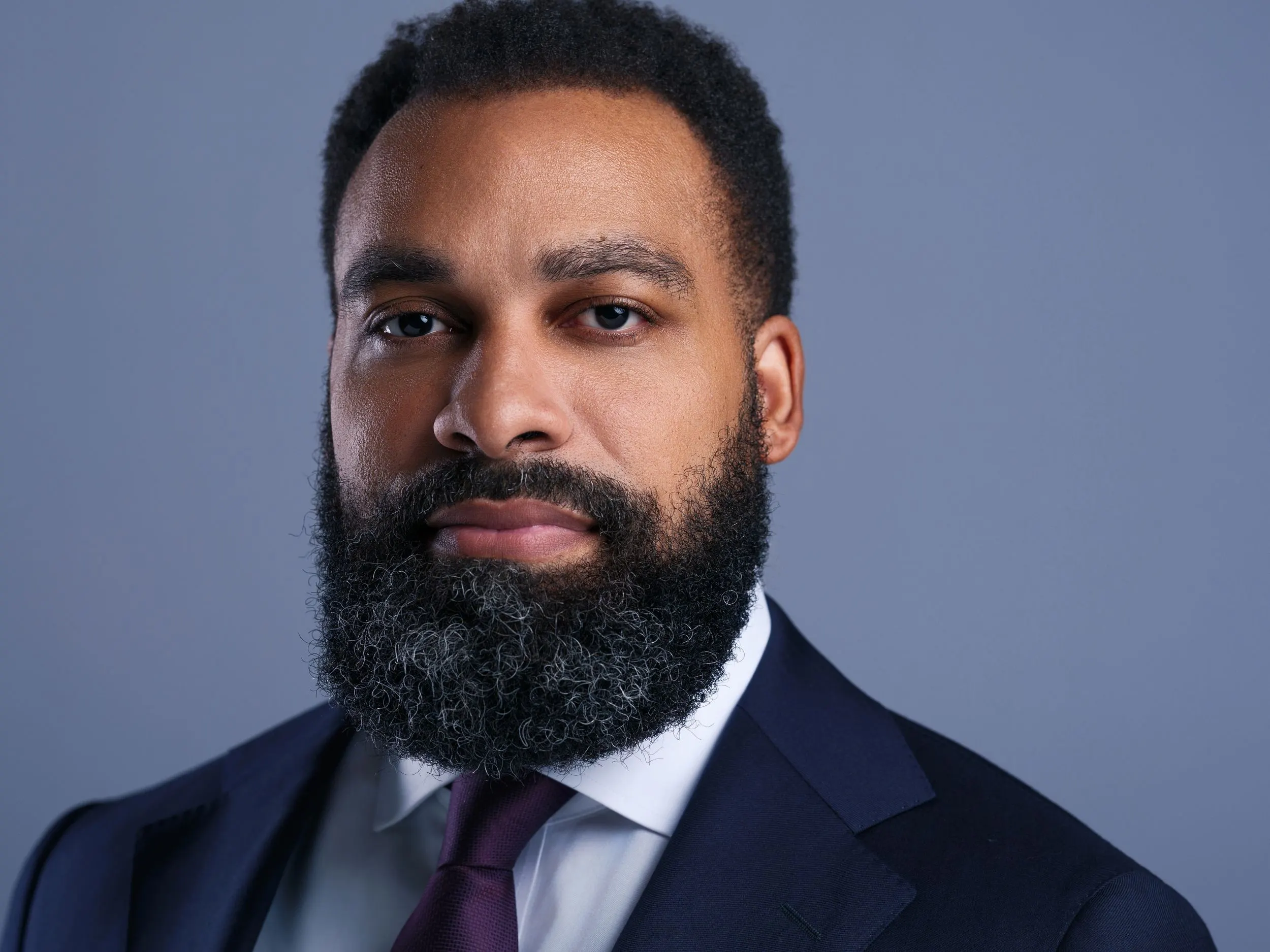 Headshot of an Egbuonu Law attorney with a full beard in a navy suit and burgundy tie on a gray backdrop