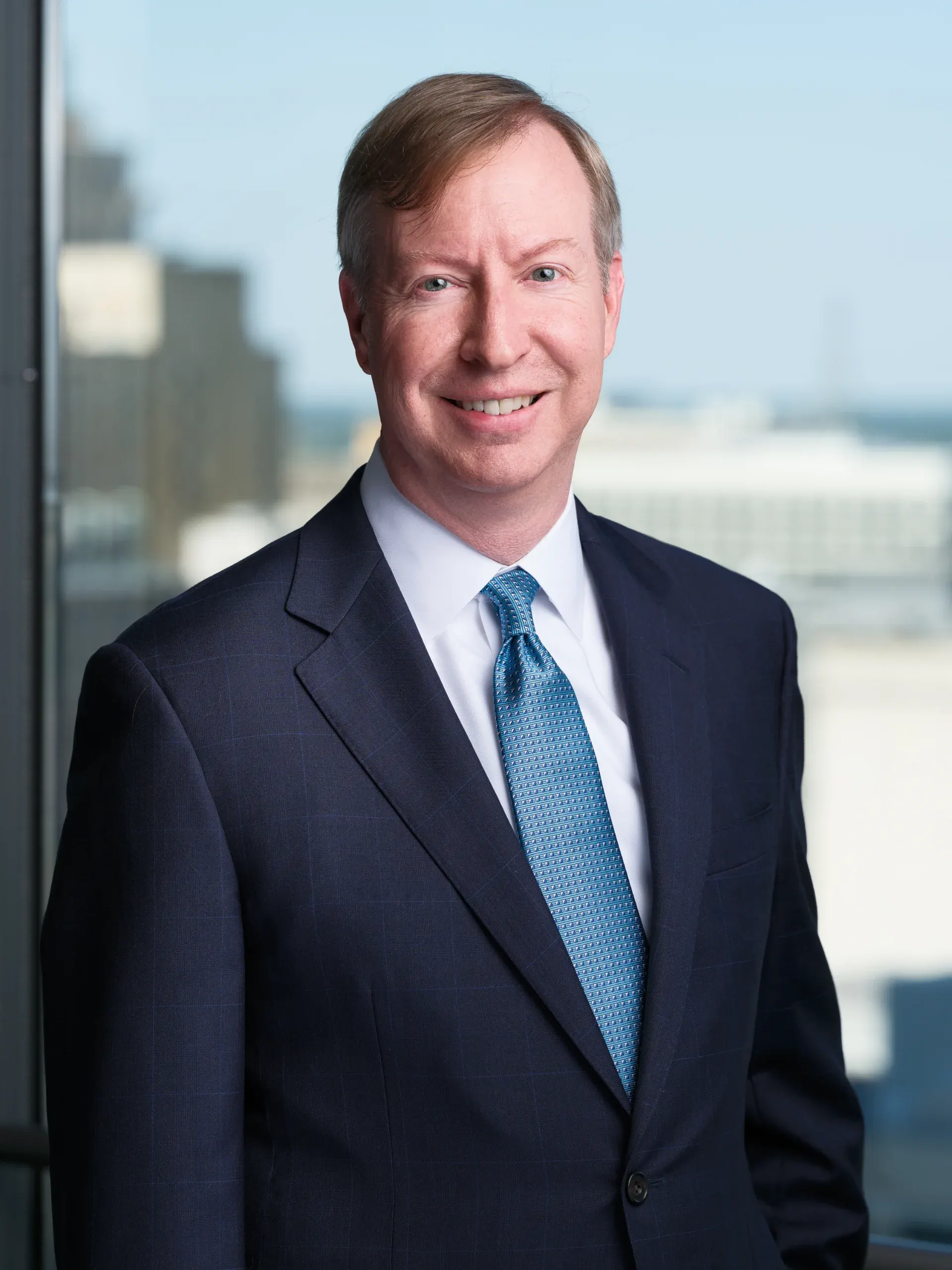 Executive headshot of a man in a navy suit and light blue tie beside a window over downtown Houston