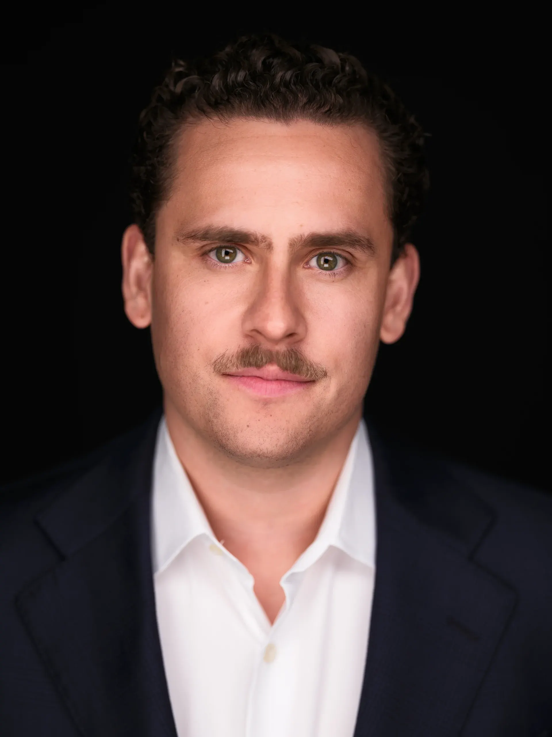 Houston headshot of a man with a mustache in a dark blazer and white open-collar shirt on a black backdrop
