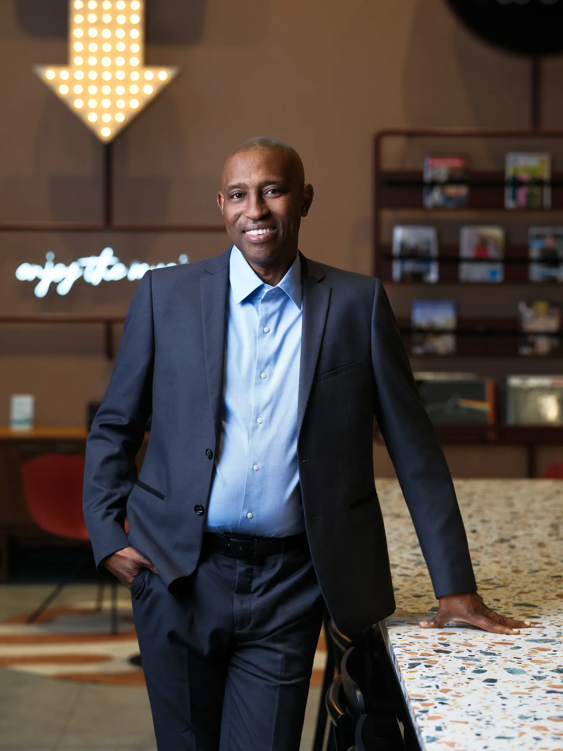 Executive portrait of a man in a navy suit leaning on a terrazzo counter in a Houston lobby with a neon sign