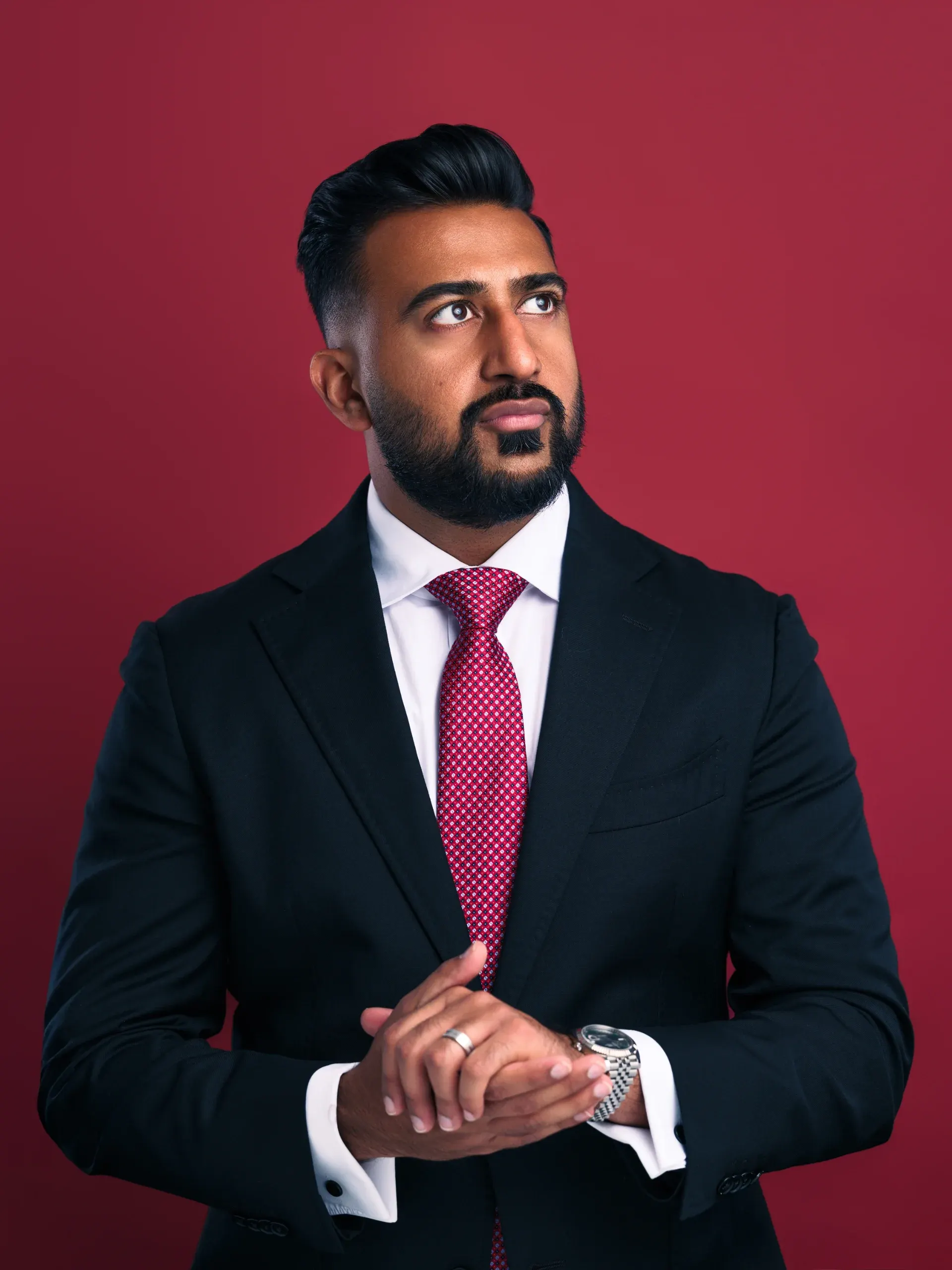 Houston headshot of a bearded man in a black suit and pink tie, hands clasped, on a red backdrop