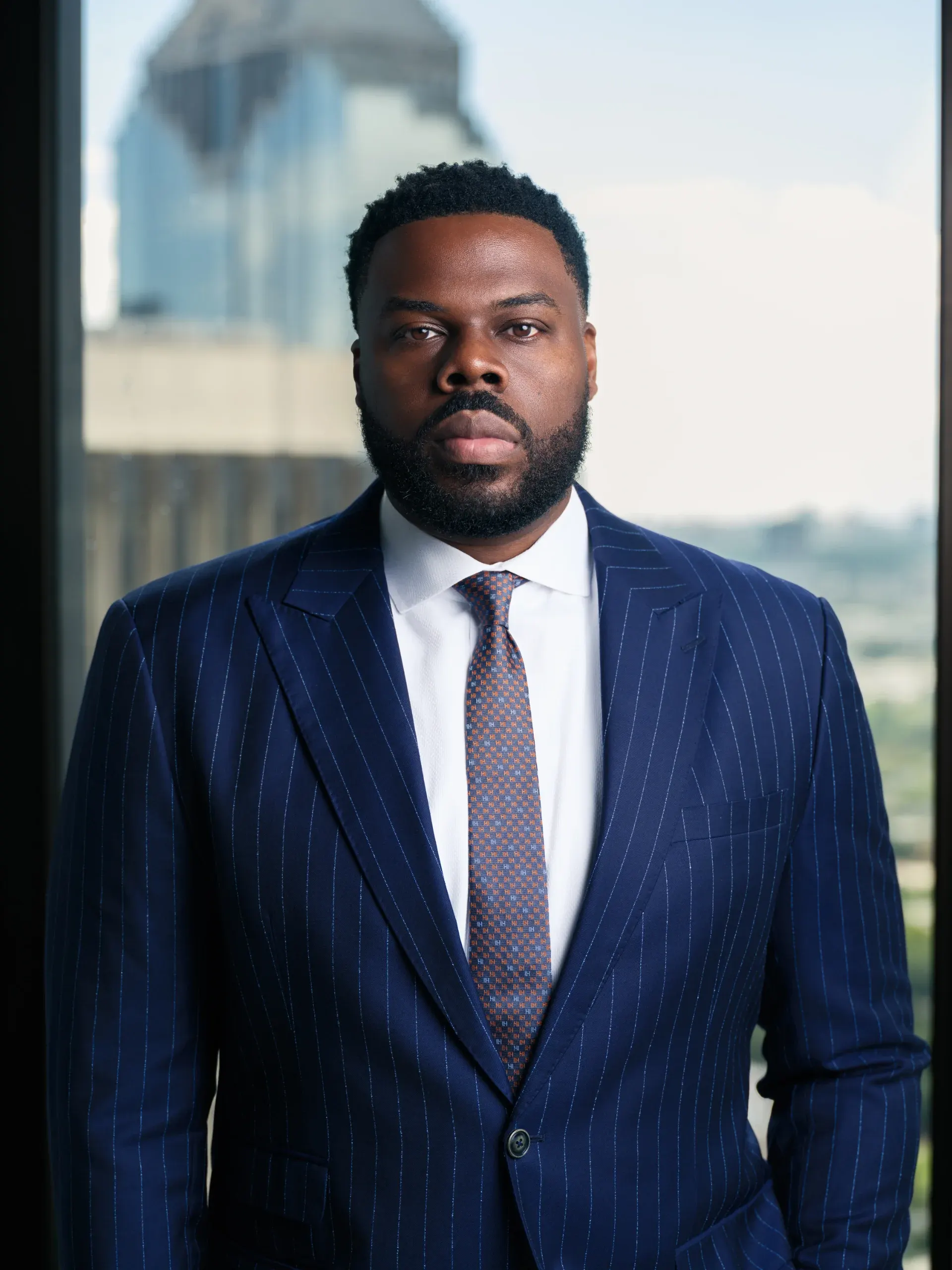 Executive portrait of a man in a navy pinstripe suit beside a high-rise window over Houston