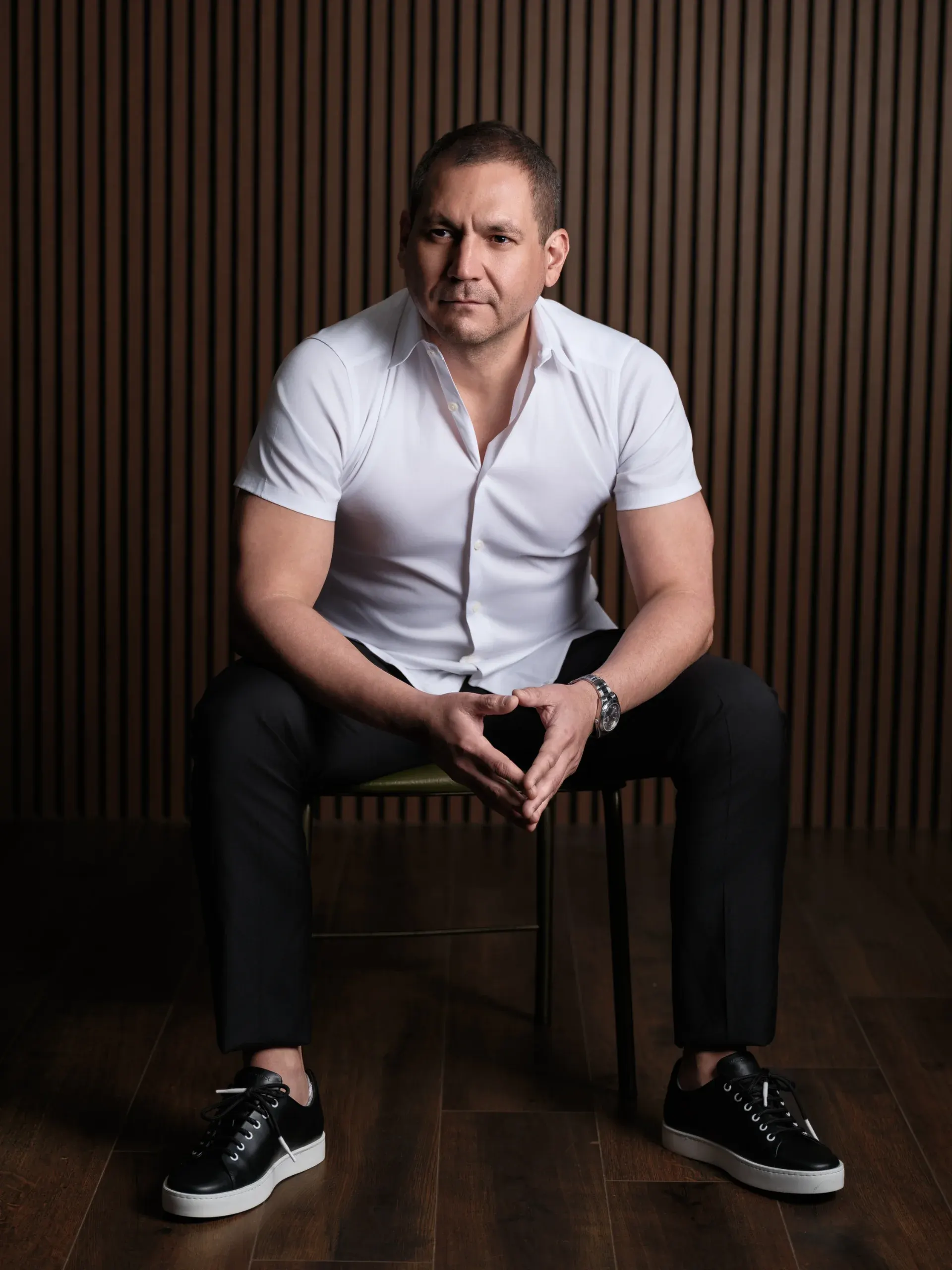 Portrait of a man in a white shirt seated on a chair in front of a dark wood slat wall