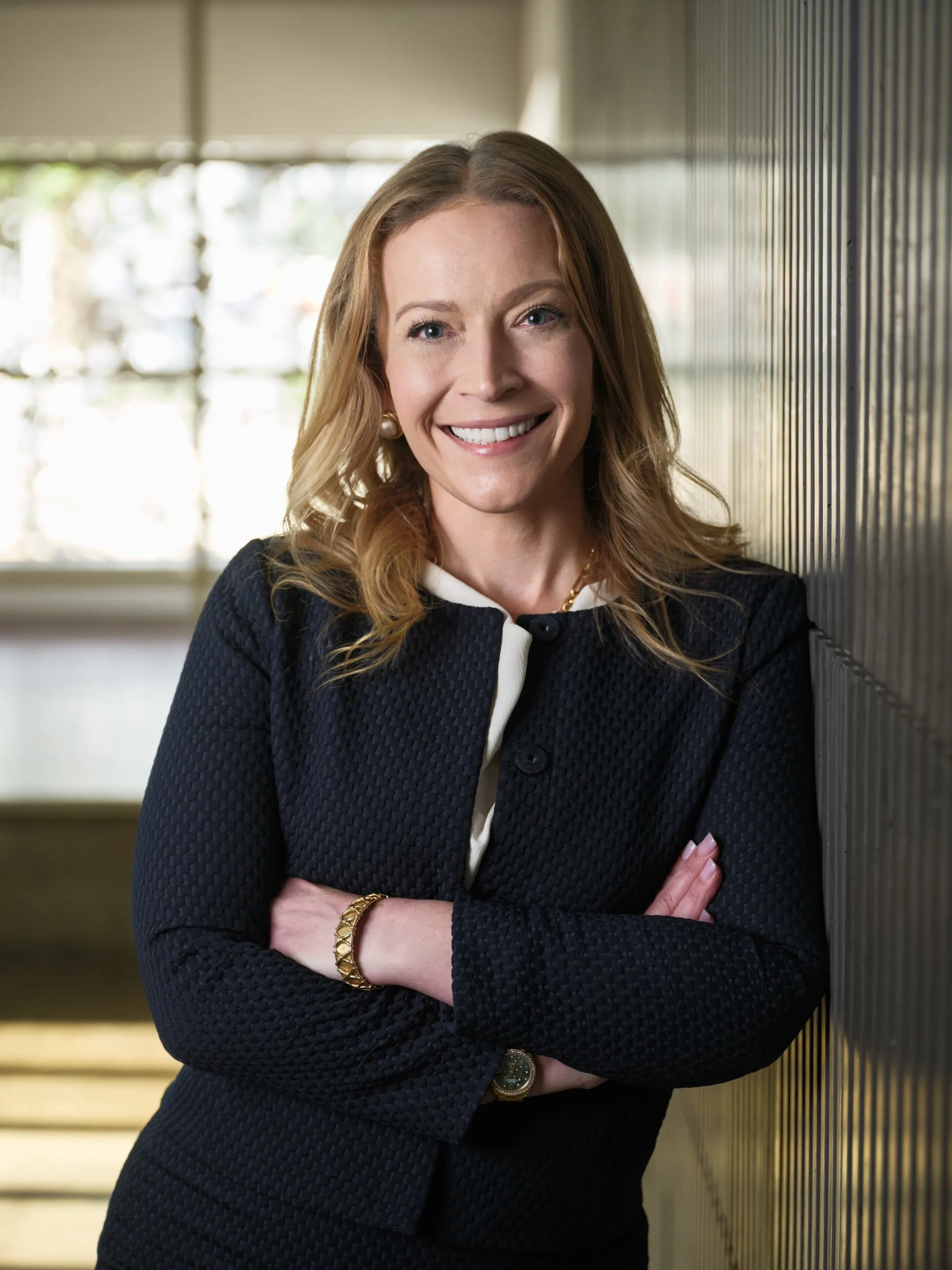 Houston attorney portrait of a smiling woman in a navy suit, arms crossed, leaning on a slatted wall by a window