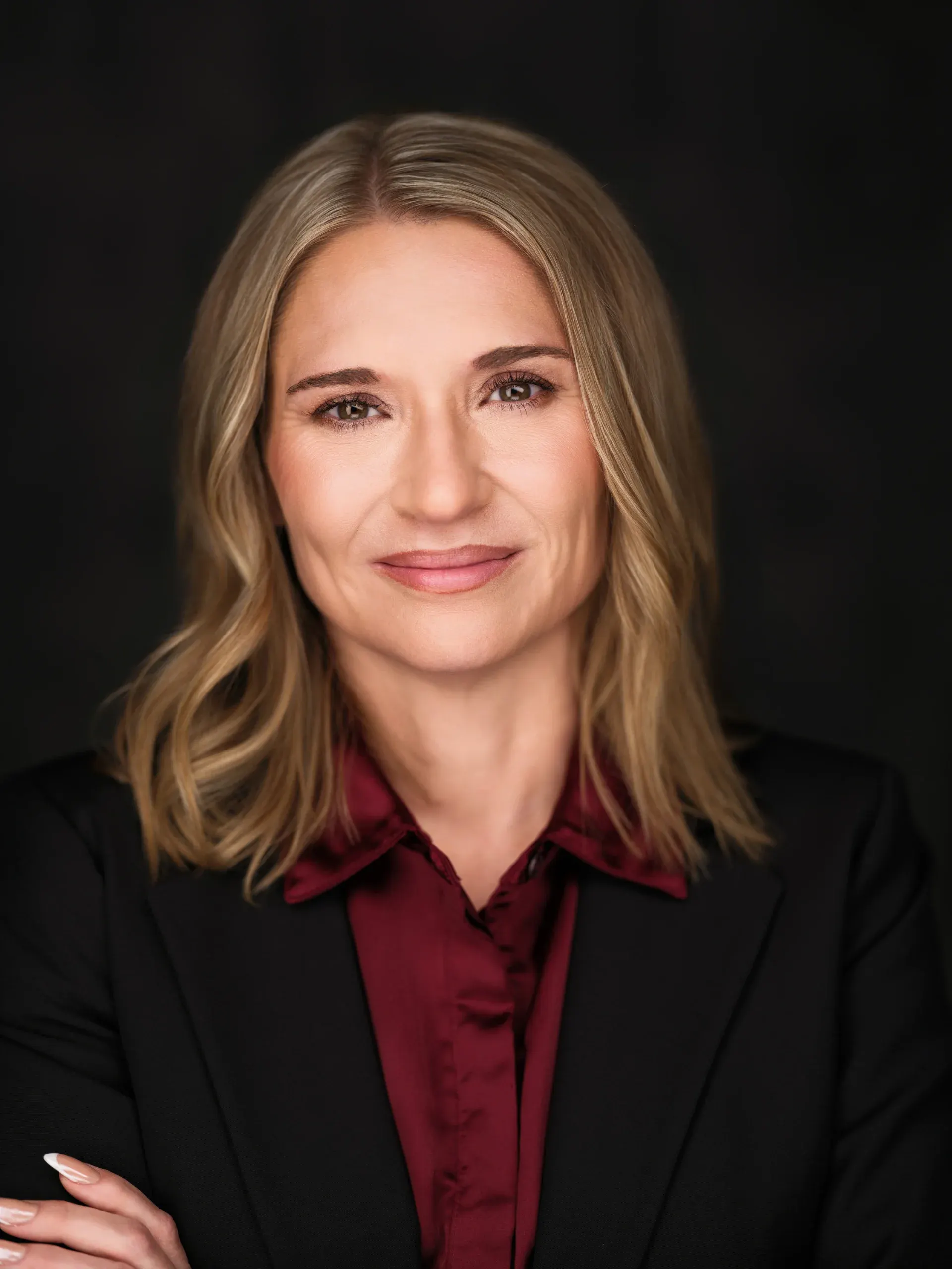 Houston headshot of a woman in a black blazer and burgundy blouse, arms crossed, on a black studio backdrop