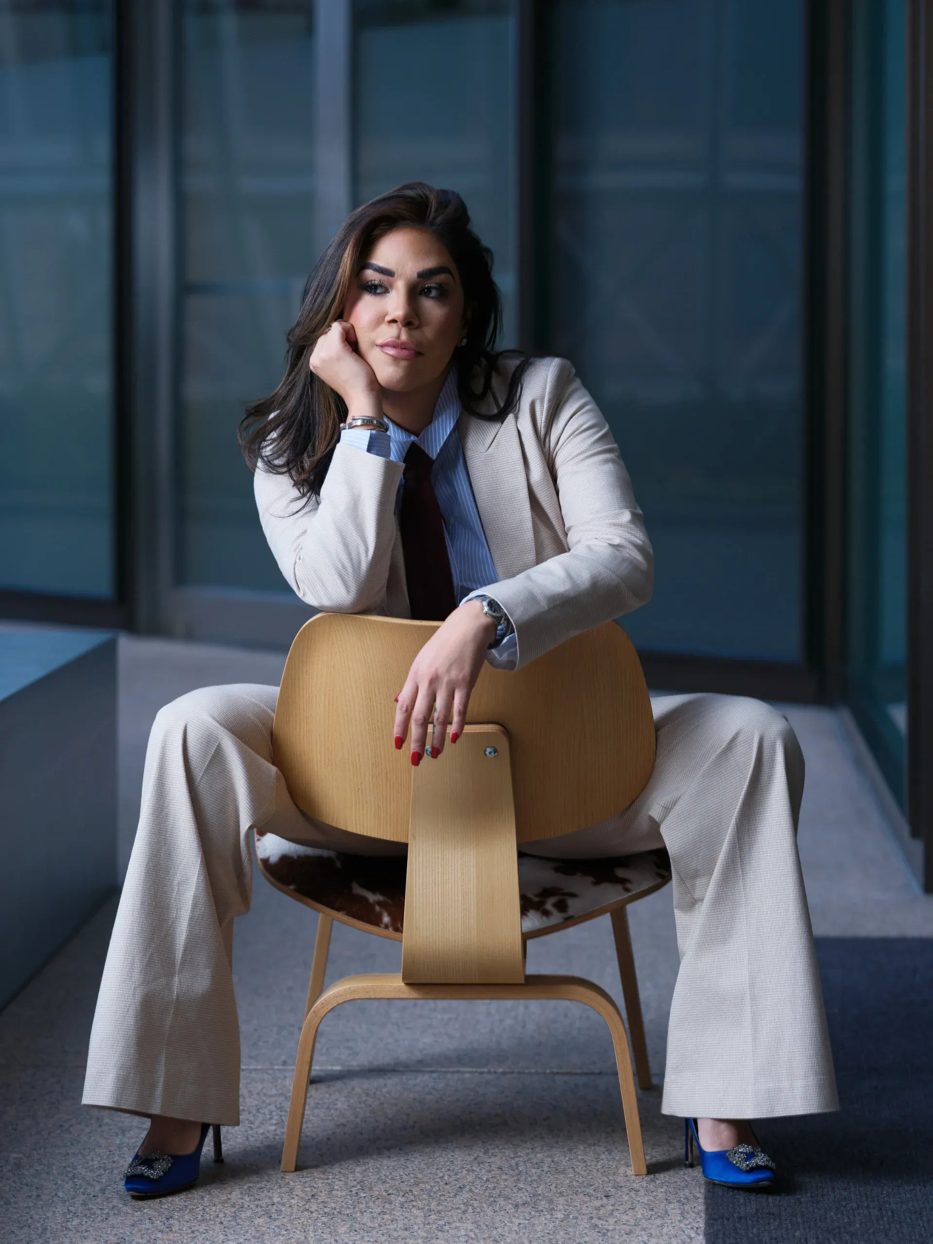 Personal branding portrait of a woman in a cream suit seated on a molded plywood chair beside a glass wall