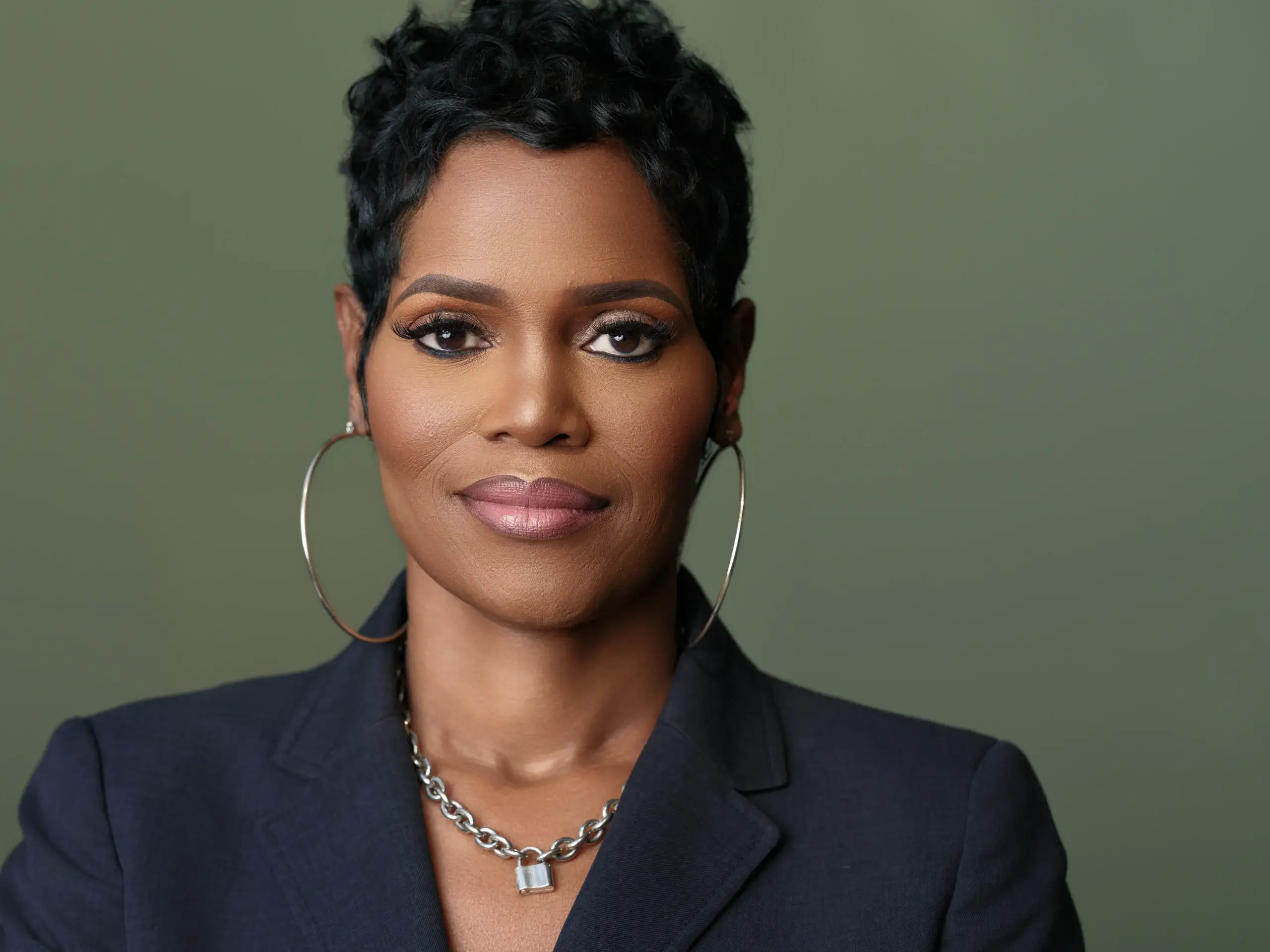 Houston headshot of a woman with short dark hair, hoop earrings, and a dark blazer on an olive green backdrop